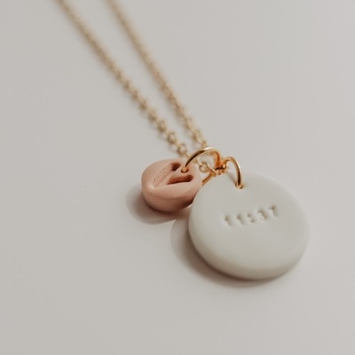 Colar dourado com dois pendentes cerâmicos, um branco redondo com inscrições, outro rosa em formato de nó.