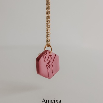 Colar com pendente hexagonal rosa e corrente dourada, fundo claro, texto Ameixa