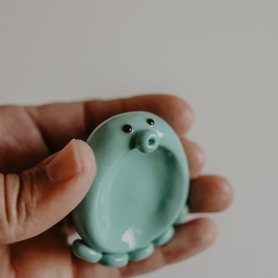 Objeto cerâmico verde azulado com formato arredondado e olhos pretos nas mãos.