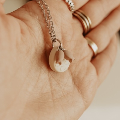 Colar prateado com pendente branco e duas pequenas figuras na mão de pessoa com anéis