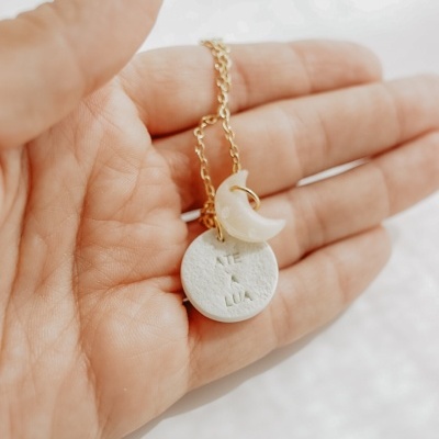 Colar dourado com dois pendentes, um redondo branco com texto e uma lua crescente perolada