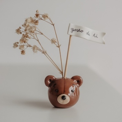 Vaso castanho em forma de cabeça de urso com flores secas e bandeira branca com texto