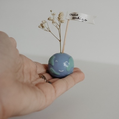 Mini vaso cerâmico planeta Terra com flores secas e bandeira 'cresço contigo' numa mão