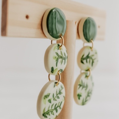 Brincos com discos de cerâmica com padrão de folhas verdes, pendurados numa estrutura de madeira.