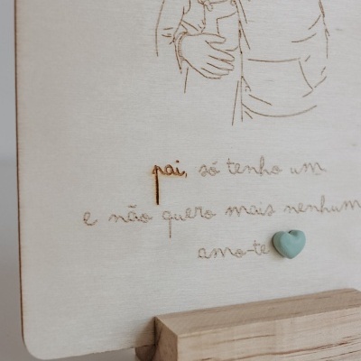 Placa de madeira clara gravada a laser com ilustração de duas pessoas e texto em português, apoiada numa base de madeira.