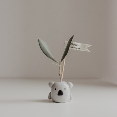 Vaso branco em cerâmica com formato de coala, raminhos verdes e bandeira branca com texto