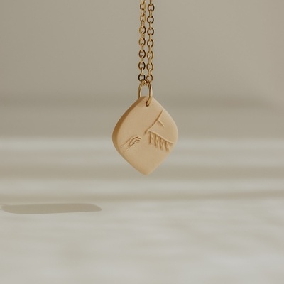 Colar dourado com pendente bege em forma de losango com relevo de mão