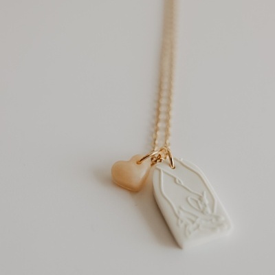 Colar dourado com dois pendentes, um coração e um retângulo branco com padrão abstrato