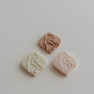 Três pendentes quadrados em tons terra com relevo de figura feminina e texto Modelo 1