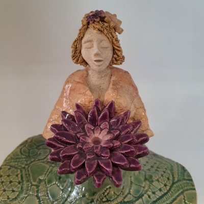 Figura decorativa de mulher em cerâmica segurando flor roxa sobre superfície verde com textura