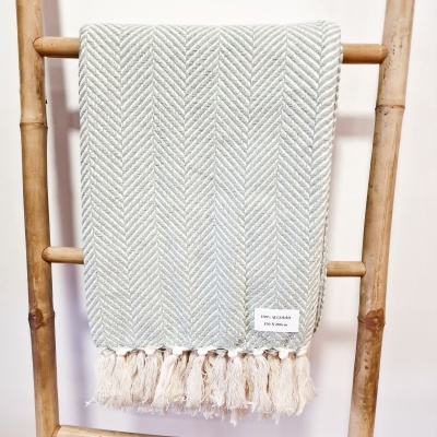 Manta cinza claro com padrão em espinha de peixe e franjas pendurada numa escada de madeira.