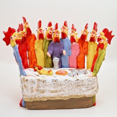 Figuras cerâmicas coloridas de galinhas e galos em volta de uma mesa com toalha branca