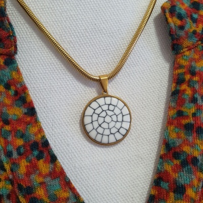 Colar dourado com pendente redondo branco e preto sobre tecido branco com tecido colorido ao lado