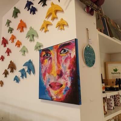 Quadro abstrato colorido de rosto humano na parede com esculturas de pássaros coloridos ao redor e prateleira com produtos