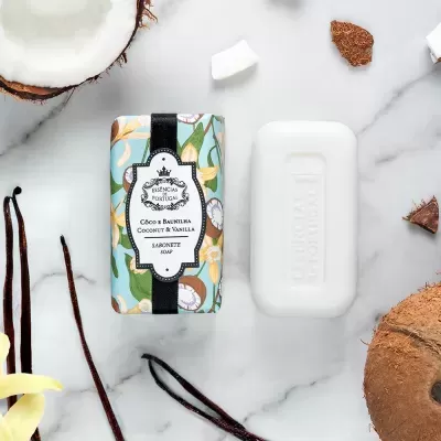 Sabonete branco com embalagem decorativa floral e texto em português e inglês, rodeado por coco e baunilha.