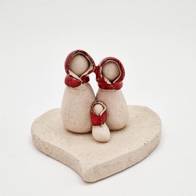 Escultura em cerâmica com duas figuras adultas e uma criança com capuzes vermelhos numa base em forma de coração.