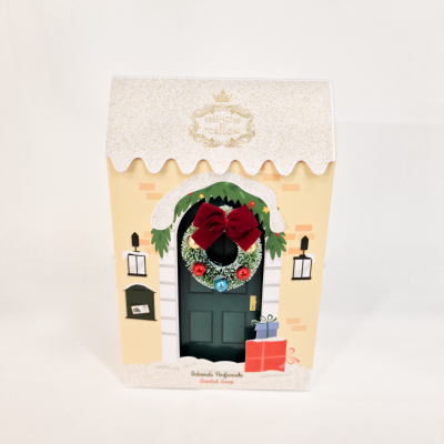Embalagem em forma de casa com porta verde, guirlanda e laço vermelho.