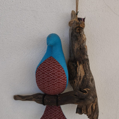 Pássaro decorativo em tecido azul e vermelho sentado em ramo de madeira pendurado numa parede branca