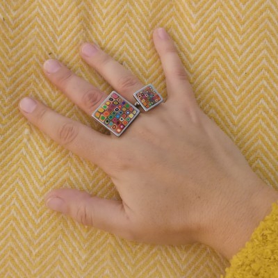 Mão com dois anéis quadrados coloridos em fundo amarelo espinha de peixe