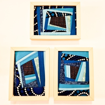 Três quadros com trabalhos têxteis em azul e preto com molduras de madeira clara