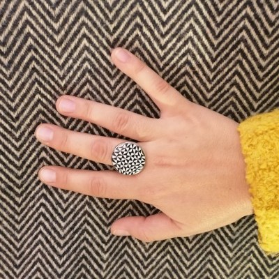Anel preto e branco com padrão geométrico triangular numa mão sobre tecido com padrão espinha de peixe.