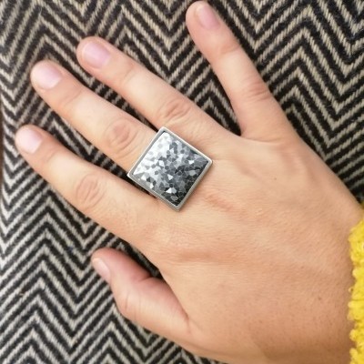 Anel quadrado prata com padrão geométrico na mão esquerda sobre fundo zig-zag preto e branco