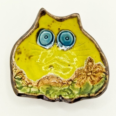 Escultura cerâmica amarela com olhos azuis e detalhes verdes e castanhos