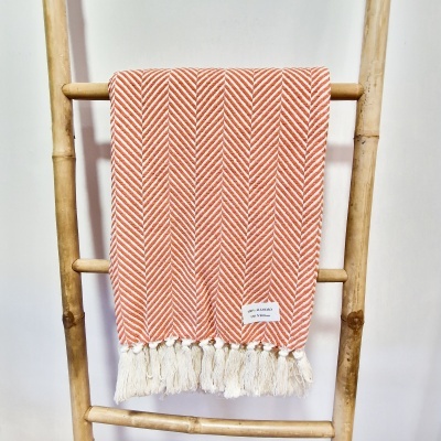 Manta laranja com padrão espinha de peixe e franjas brancas pendurada numa escada de bambu