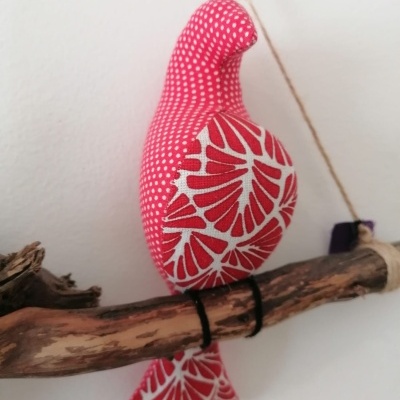 Pássaro decorativo em tecido vermelho com padrões brancos, sentado num ramo de madeira.