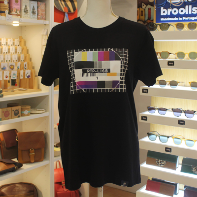 T-shirt preta com padrão gráfico colorido e texto RTP-LISB num manequim em loja de acessórios.