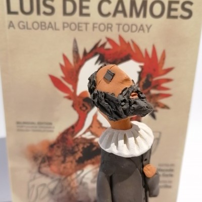 Figura decorativa de homem com barba e gola branca diante de capa de livro intitulada LUÍS DE CAMÕES A GLOBAL POET FOR TODAY.