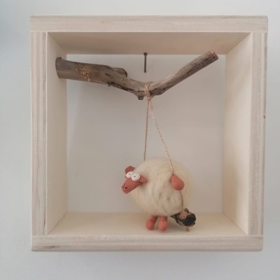 Decoração de ovelha em lã e argila pendurada num ramo dentro de caixa de madeira clara.