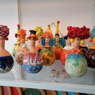 Figuras decorativas de cerâmica coloridas com cabeças de boneca e acessórios de tecido, expostas em prateleira