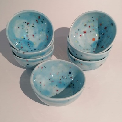 Seis tigelas cerâmicas azuis claras com manchas coloridas, empilhadas e dispostas sobre superfície branca.