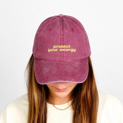 Pink baseball cap with yellow protect your energy embroidery on a woman wearing white and a gold chain