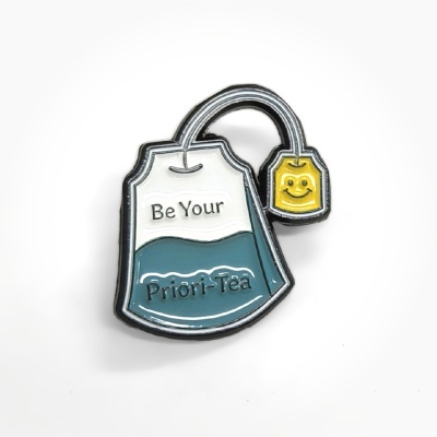 Enamel pin shaped like a tea bag with text Be Your Priori-Tea and smiling yellow tag