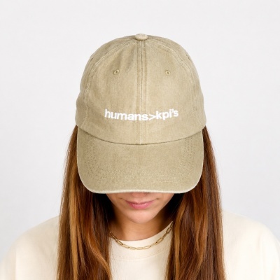 Beige baseball cap with white text humans>kpis worn by person with long brown hair