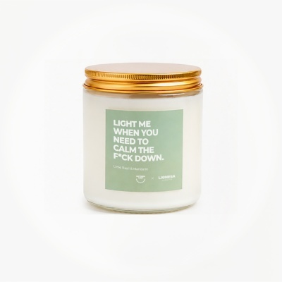 White candle in glass jar with gold lid and green label with calming text