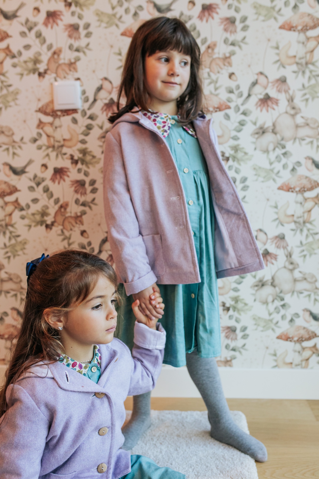Casaco Bosco Duas meninas com casacos lilás e vestidos verdes com gola floral num quarto com papel de parede floral.