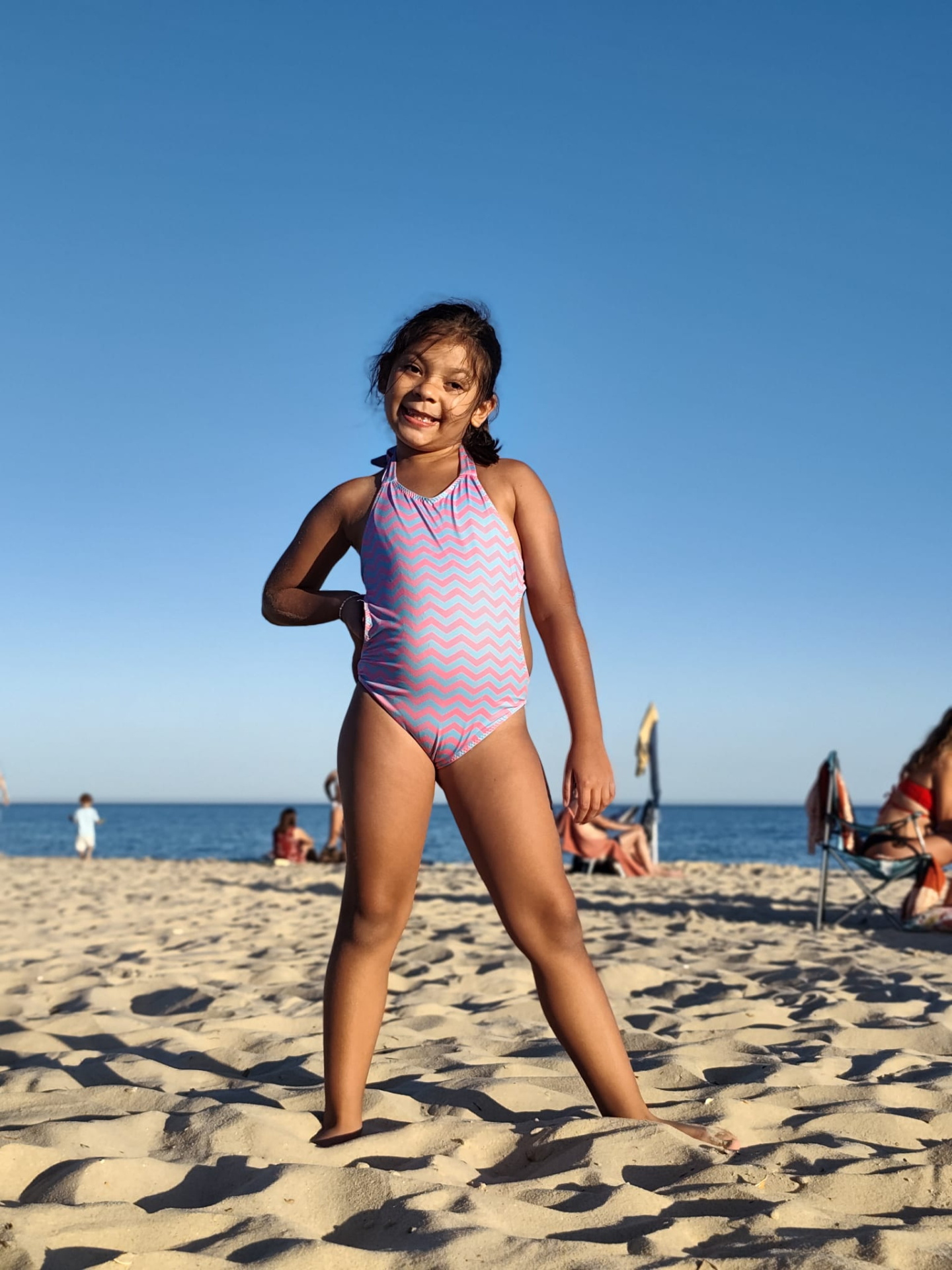 Trikini Blue Waves Menina de fato-de-banho cor-de-rosa com padrão ondulado azul na praia sob céu azul.