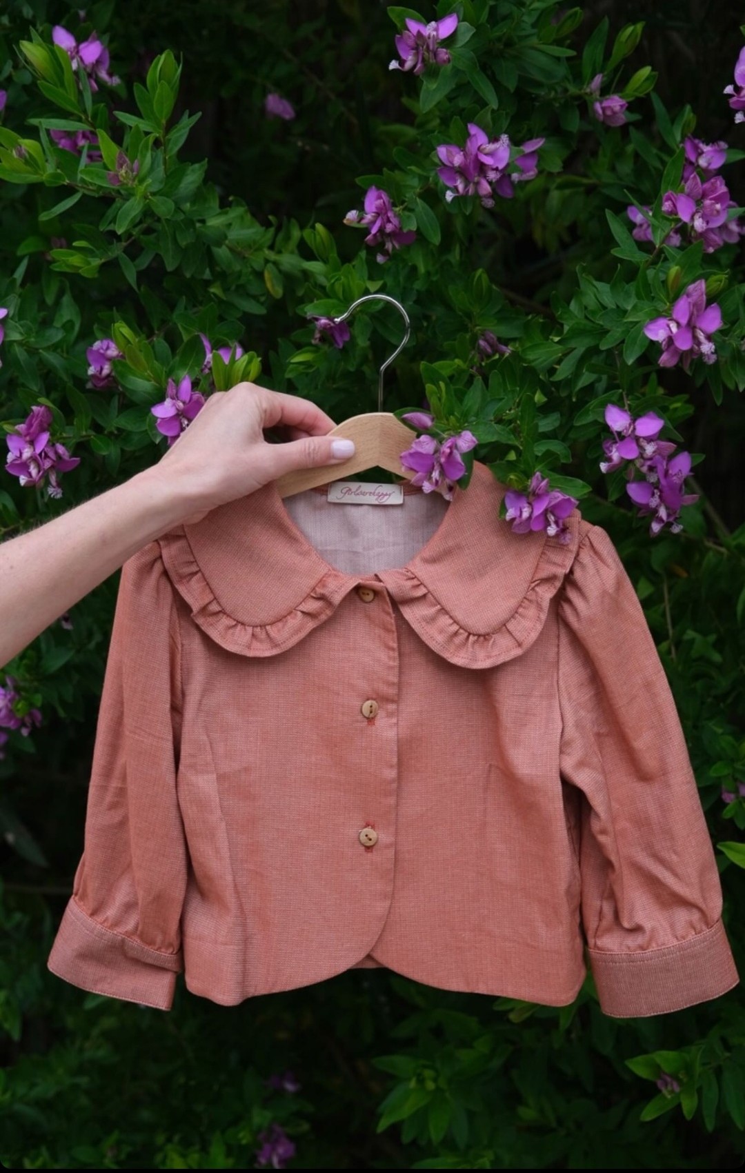 Camisa Telha Blusa cor de rosa para criança com gola grande e folhos num cabide segurado por uma mão com fundo de folhas verdes e flores roxas