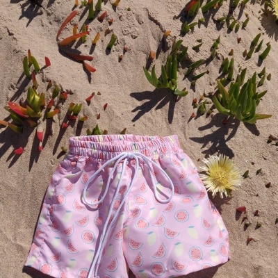 Calções de banho rosa com padrão de melancia na areia com plantas verdes e flores amarelas