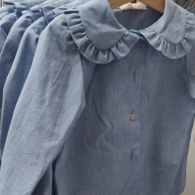 Camisas azul claro para crianças com gola folhada penduradas em cabides de madeira num carrinho de roupa branco