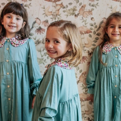 Três meninas com vestidos verdes e golas florais num ambiente com papel de parede com padrão floral.