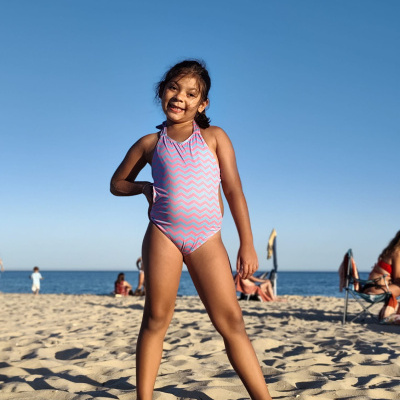 Menina de fato-de-banho cor-de-rosa com padrão ondulado azul na praia sob céu azul.