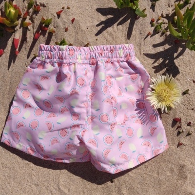 Calções de banho rosa com padrão de melancias na areia, com vegetação verde e flor amarela próxima.