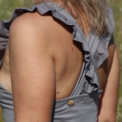 Alças largas com folhos e botões numa roupa cinzenta, vista das costas com fundo natural.