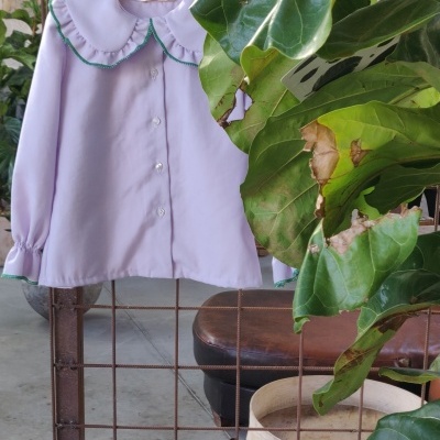 Camisa infantil rosa clara com gola redonda pendurada numa grade metálica com plantas verdes ao redor