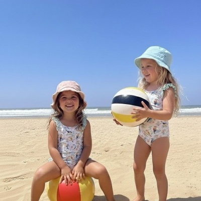Duas meninas na praia com fato de banho floral e chapéus, brincando com bolas coloridas