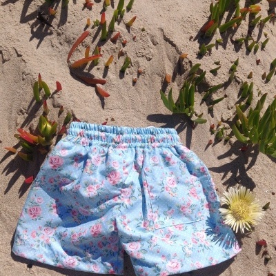 Calções de praia azul claro com padrão floral sobre areia com plantas e flores amarelas